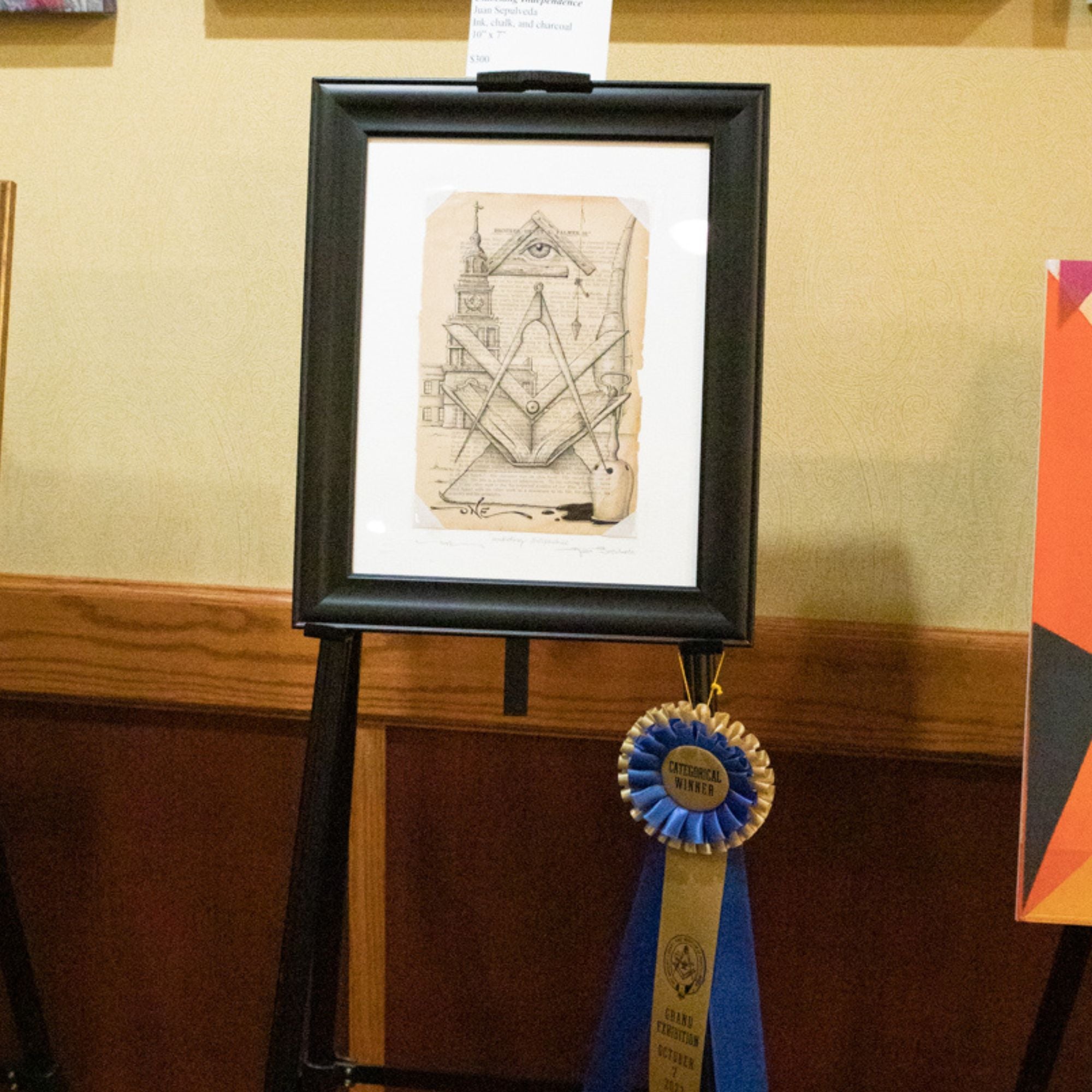A framed artwork titled 'Unlocking Independence' displayed on an easel, featuring ink, chalk, and charcoal on a white page, with a blue ribbon award in front of it.