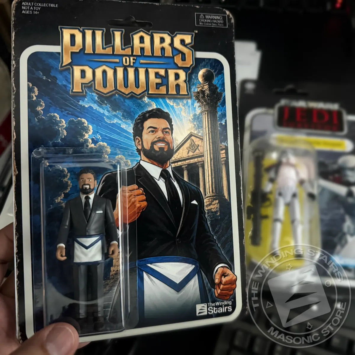 A packaged action figure of 'Pillars of Power' from The Walking Stairs, held in a hand. The figure is dressed in a black suit with a white apron.